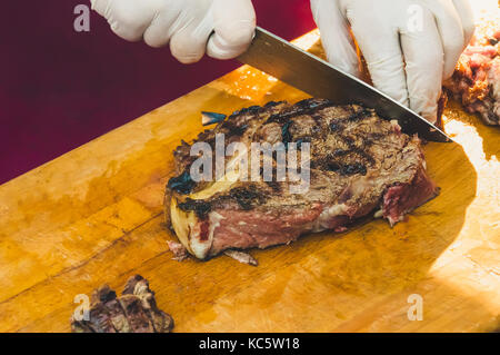 Male chef cutting big pièce de boeuf sur planche de bois dans une cuisine de restaurant. la préparation faire cuire l'homme steak et garniture de légumes. mains cuisiniers dans propre blanc Banque D'Images