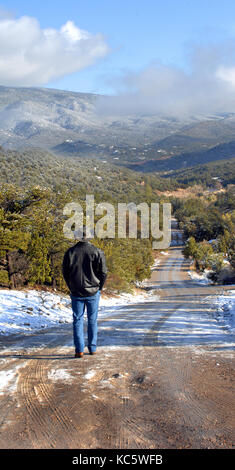 L'homme, vêtue de jeans et manteau noir, marche dans une route enneigée à tijeras, Nouveau Mexique. gravier disparaît dans la distance et la sandia mountai Banque D'Images