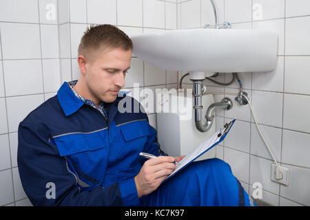 Jeune plombier writing on clipboard avec stylo dans la cuisine prix Banque D'Images