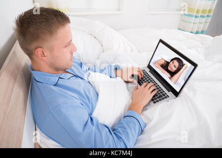 Jeune homme assis sur le lit le chat vidéo avec femme Banque D'Images