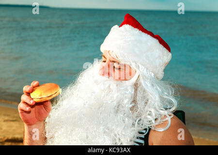 Père Noël sur la plage de manger un hamburger. Le concept de l'alimentation malsaine. Banque D'Images