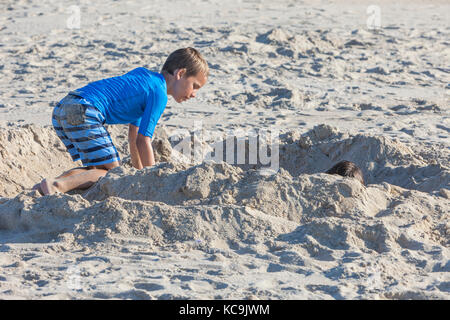 Avon, Outer Banks, Caroline du Nord, USA. Jeune garçon contrôle sur son frère, enterré dans le sable. Banque D'Images