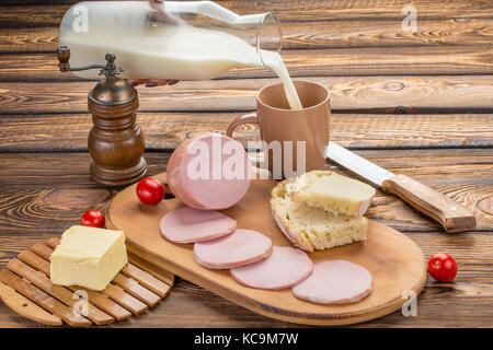 Still Life de tranches de saucisse à cuire avec des ingrédients. petit-déjeuner rural - saucisse à cuire, pain, lait, beurre, tomates sur fond de bois. le lait Banque D'Images