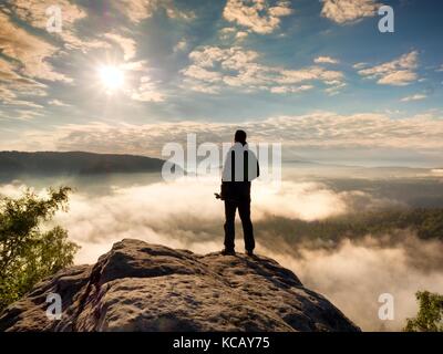 Photographe professionnel avec trépied dans les mains sur la falaise et la pensée. fogy, paysage de rêve soleil misty blue dans une belle vallée. Banque D'Images