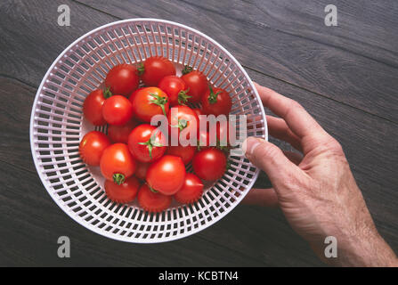Un homme part ramasser une passoire rouge vif de tomates mûres sur une surface de travail en bois, plan de travail dans une cuisine. Banque D'Images