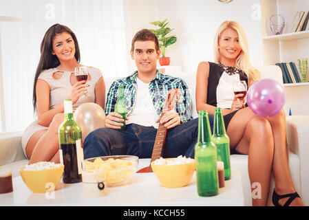 Trois jeunes gens sourire à l'accueil partie. Ils s'asseoir et boire du vin et de la bière. En regardant la caméra. Banque D'Images