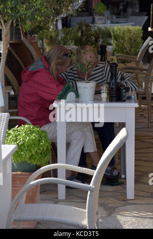 Un aîné ou d'un couple plus âgé à la recherche, à l'aide du menu dans une taverne grecque de s'asseoir à une table et des chaises sous un arbre et de parler les uns aux autres. Banque D'Images