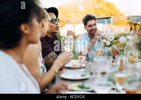 Jeune homme de rire alors qu'il était assis avec des amis à l'extérieur partie. Les amis de l'été profiter de repas au restaurant en plein air. Banque D'Images
