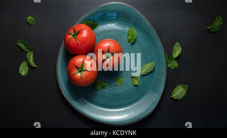 Tomates mûres et de feuilles de menthe dans une assiette. Les tomates dans une plaque en céramique sur fond noir. Banque D'Images