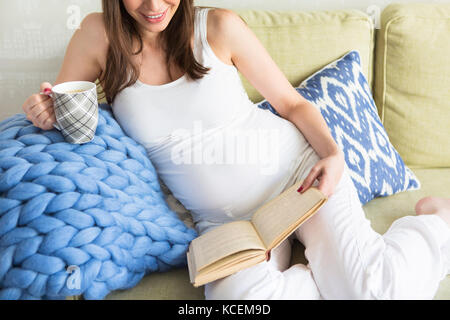 Jolie jeune femme enceinte à l'intérieur assis sur un canapé et de la lecture Banque D'Images