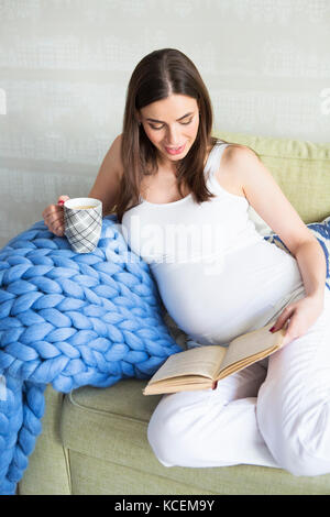 Jolie jeune femme enceinte à l'intérieur assis sur un canapé et de la lecture Banque D'Images