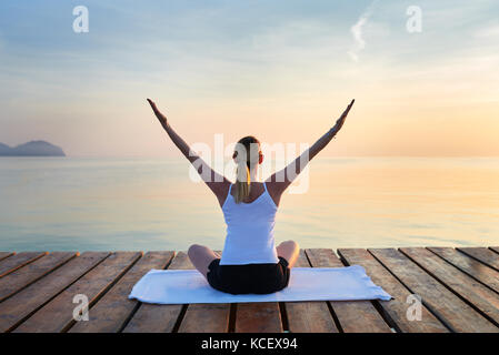 Vue arrière du jeune femme était assise sur la pratique du yoga serviette par mer au coucher du soleil Banque D'Images