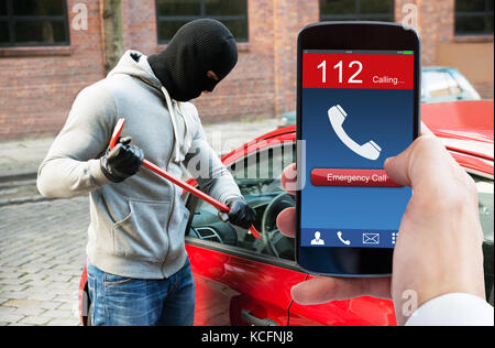 La main de la personne à l'appel d'urgence sur téléphone mobile burglar opening car door with crowbar Banque D'Images