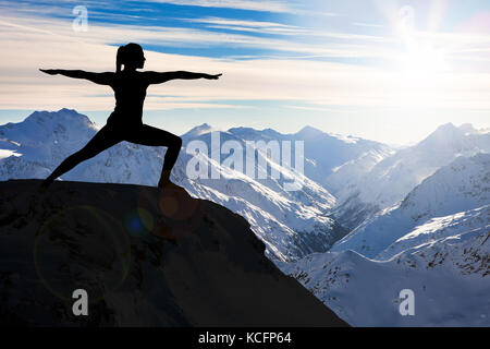 Silhouette d'une femme pratiquant le yoga sur une falaise contre les montagnes enneigées Banque D'Images