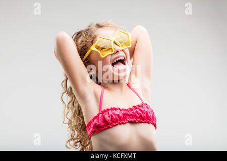 Relations sérieuses in vive little girl wearing trendy lunettes avec lentilles jaune en forme de cœur à rire comme elle tient ses bras derrière sa tête et long curly blonde h Banque D'Images