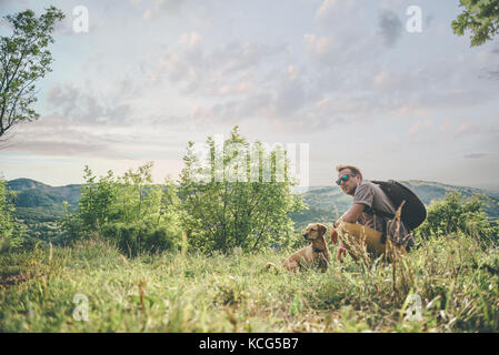 L'homme portant des lunettes de soleil et son sac à dos posé sur le pré avec un petit chien jaune Banque D'Images