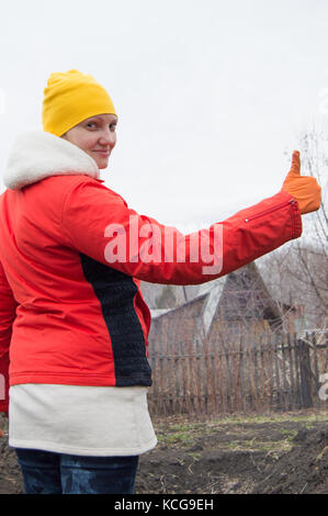 Young smiling woman jardinier dans un chapeau et une veste avec pouce vers le haut. Banque D'Images