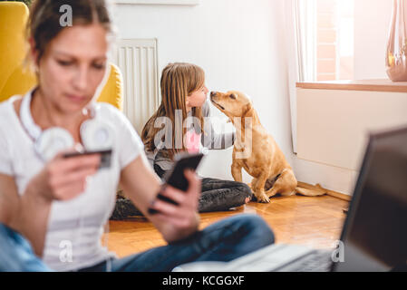 Petite fille jouant avec son chien à la maison mère alors que les achats en ligne avec smart phone Banque D'Images