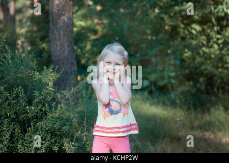 Peu de belle fille blonde debout dans les bois. elle rit elle les mains jointes Banque D'Images