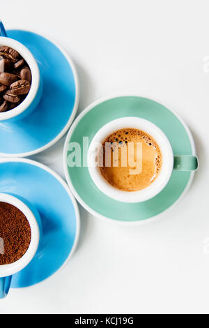 Trois tasses de café : café moulu (poudre), les grains de café et espresso fraîchement préparés dans des tasses en céramique colorée (bleu et vert menthe) sur le livre blanc Banque D'Images