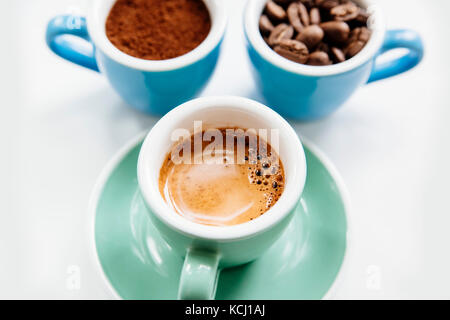 Trois tasses de café : café moulu (poudre), les grains de café et espresso fraîchement préparés dans des tasses en céramique colorée (bleu et vert menthe) sur le livre blanc Banque D'Images