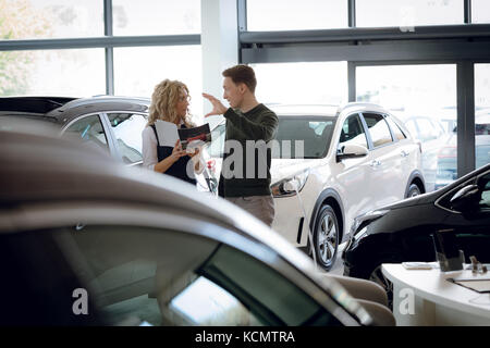 La clientèle masculine des gestes tout en discutant avec saleswoman in car showroom Banque D'Images