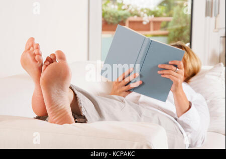 La femme est allongée sur le canapé, a ses pieds et est en train de lire un livre. Banque D'Images