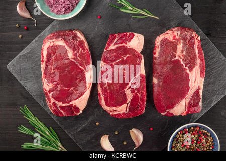 Un gros plan de trois tranches de viande crue, filet de boeuf, en plongée des textures sur noir avec des branches de romarin, sel, poivre et d'ail Banque D'Images