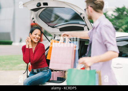 Man shopping donne à asian woman sitting on car. accro, amour, couple, multiethniques ou occasionnels concept de vie Banque D'Images