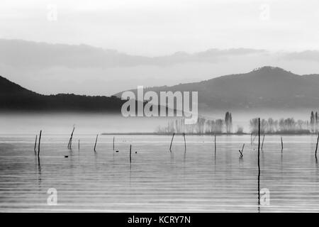 Un lac au crépuscule, avec des tons doux, des collines et montagnes, et quelques plantes et poteaux en bois au milieu de la brume Banque D'Images