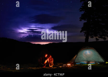 Nuit en camping dans les montagnes. Couple de touristes ont un reste à un feu allumé sous la tente près de l'incroyable nuit ciel plein d'étoiles et Milky Way. l Banque D'Images