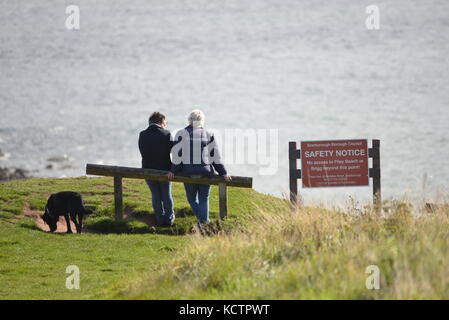 Deux personnes se sont assises à côté d'un panneau de sécurité avec leur chien qui a vue sur la mer lors d'une journée d'automne ensoleillée, Filey, dans le North Yorkshire UK Banque D'Images