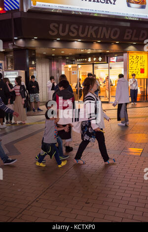 Photo de nuit de mère en marche avant du bâtiment susukino avec deux petits garçons jouant avec son pull blanc à marcher derrière elle. Banque D'Images