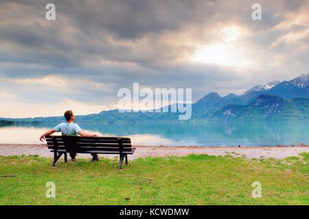 Seul l'homme est assis sur un banc à côté d'un azure Mountain Lake. man vous détendre et regarder les hauts sommets des Alpes, au-dessus du lac miroir. Banque D'Images