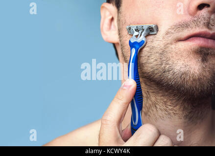Vue latérale d'un jeune homme barbu avec un rasoir brutal. La moitié de la face sur le fond bleu. Banque D'Images