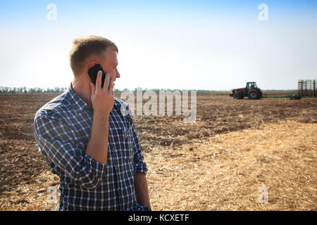 Jeune homme au téléphone dans un champ et un tracteur sur un arrière-plan. Concept de l'agriculture et des travaux publics. Banque D'Images