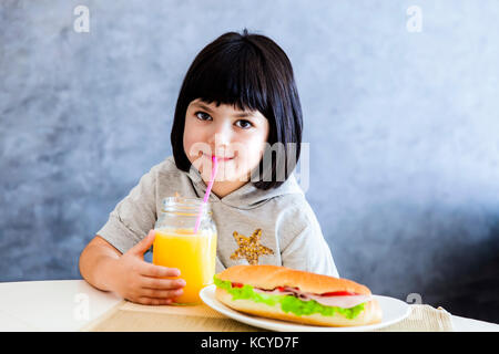 Cute little girl cheveux noir de prendre le petit déjeuner et boire le jus d'orange à la maison Banque D'Images