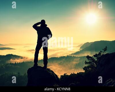 Silhouette homme haut escalade sur falaise. randonneur est montée jusqu'au pic de profiter de la vue. l'homme veille sur misty et matin brumeux en vallée de soleil du matin. Banque D'Images