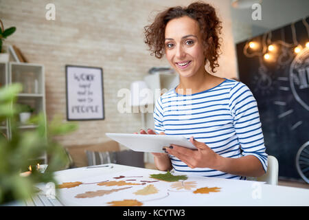 Portrait of happy young woman using digital tablet and smiling at camera tout en faisant des décorations d'automne à la maison Banque D'Images