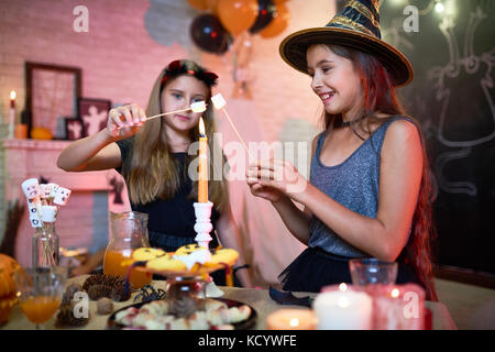 Deux amis joyeuse portant des costumes de Halloween faire frire les guimauves avec l'aide de flamme de bougie tout en appréciant home party, intérieur de décorées roo Banque D'Images