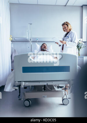 Smiling doctor with clipboard visiter homme à hôpital. Physician attending patient in hospital bed et de rapports. Banque D'Images