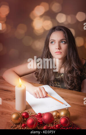 Belle jeune femme, aux yeux bleus, de penser ce qu'il faut écrire, à la lumière des chandelles, entouré de globes rouges et jaunes et des lumières de Noël. Banque D'Images