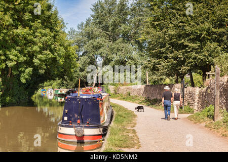 7 juillet 2017 : Bradford on Avon, Somerset, Angleterre, Royaume-Uni - couple avec un chien marchant sur le chemin de remorquage à côté d'une rangée de bateaux étroits amarrés le long des rives du t Banque D'Images