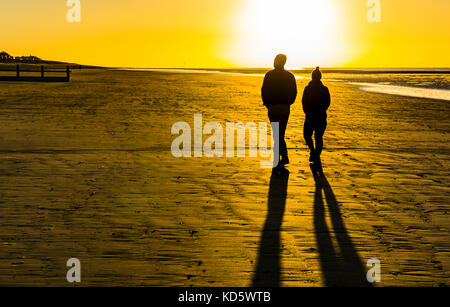 Silhouette d'un couple en train de marcher sur une plage de sable fin très tôt le matin avec de longues ombres au lever du soleil, au Royaume-Uni. Banque D'Images