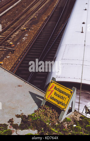 Panneau jaune avertissement de fils de contact haute tension au-dessus des rails, et blanc vitesse train dans l'arrière-plan Banque D'Images