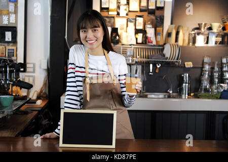 Portrait of smiling asian barista holding tasse de café avec blank chalkboard menu au comptoir dans un café. café restaurant service, la nourriture et les boissons Banque D'Images