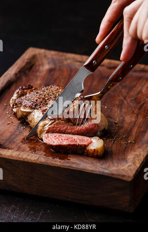 Tranches de viande grillé Steak New York faux filet avec couteau et fourchette en mains mâles sur planche de bois sur fond noir close-up Banque D'Images