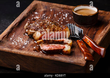 Tranches de viande grillé Steak New York faux filet avec sauce au piment et le couteau et la fourchette sur planche de bois sur fond noir Banque D'Images