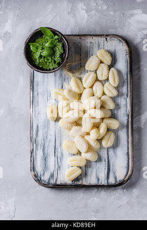 Gnocchi de pommes de terre non cuites brutes sur planche en bois blanc avec des ingrédients. La farine, sauce pesto au basilic sur fond de béton gris. Vue supérieure avec spa Banque D'Images