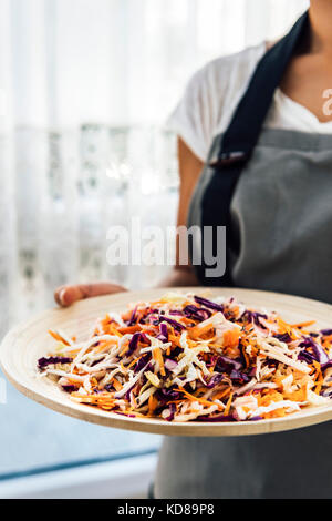 Une femme avec un T-shirt blanc et un tablier gris tenant un bol en bois avec la salade de chou faite avec des carottes râpées, chou blanc et violet photographié fro Banque D'Images